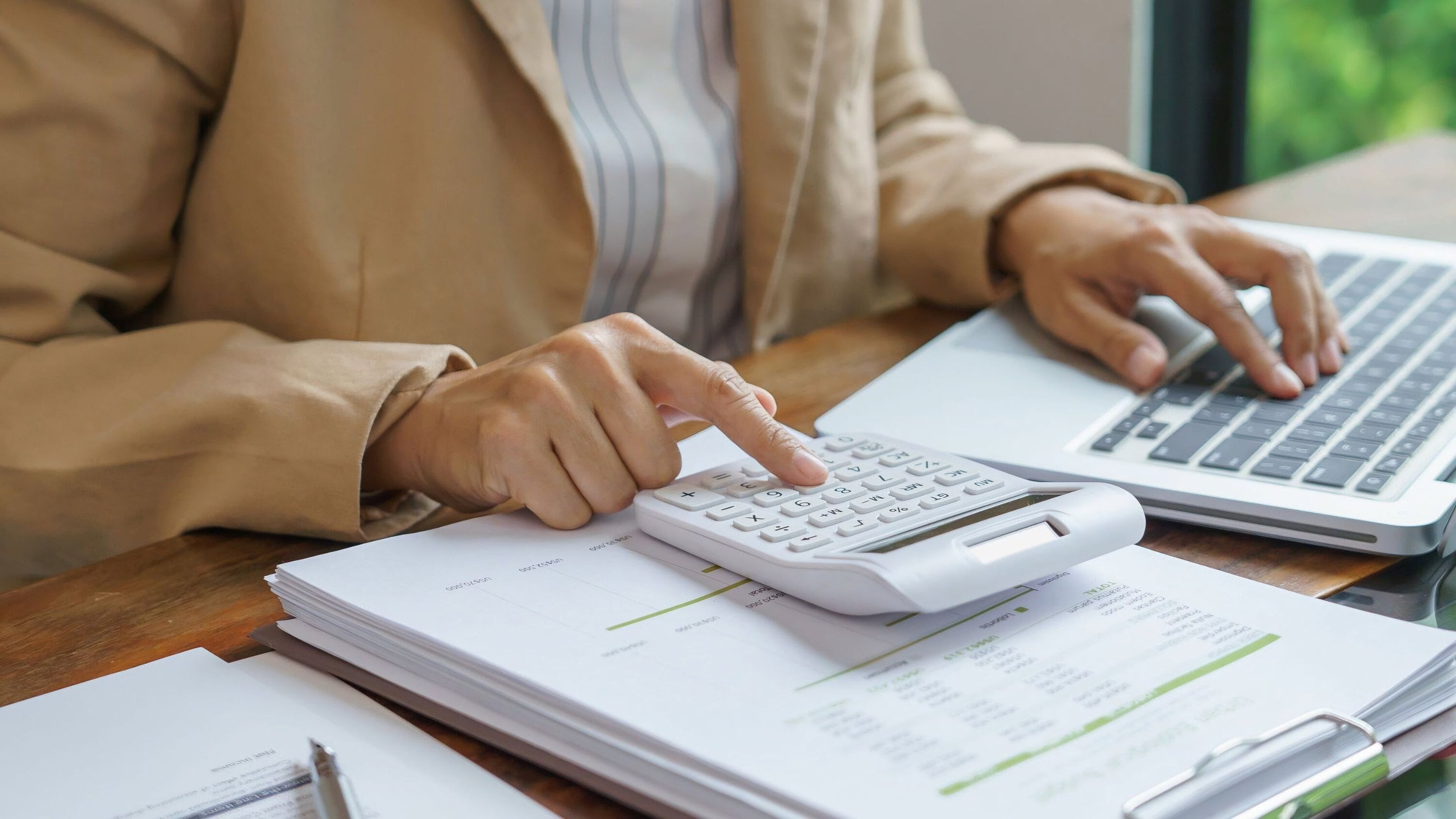Una persona trabajando en una laptop y utilizando una calculadora. Sobre el escritorio hay varios documentos y papeles, lo que sugiere que la persona está realizando tareas de oficina o contabilidad. La imagen tiene un enfoque en las manos y los elementos, sin mostrar su rostro. El entorno parece ser una oficina.