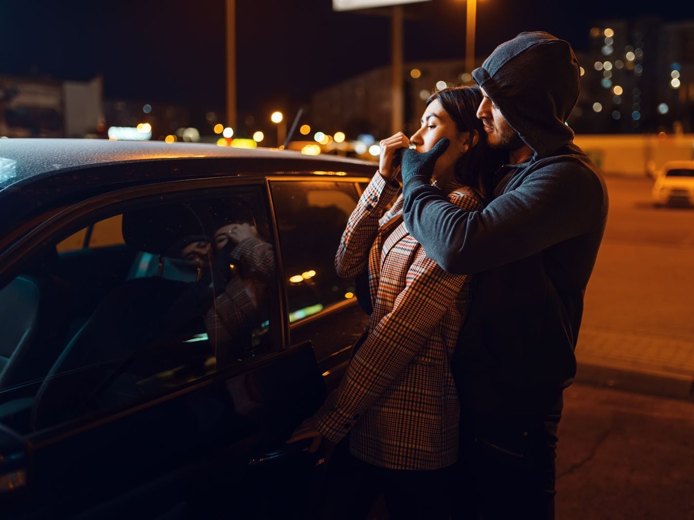 Foto ilustrativa de un hombre encapuchado amordazando con su mano a una mujer afuera de su carro