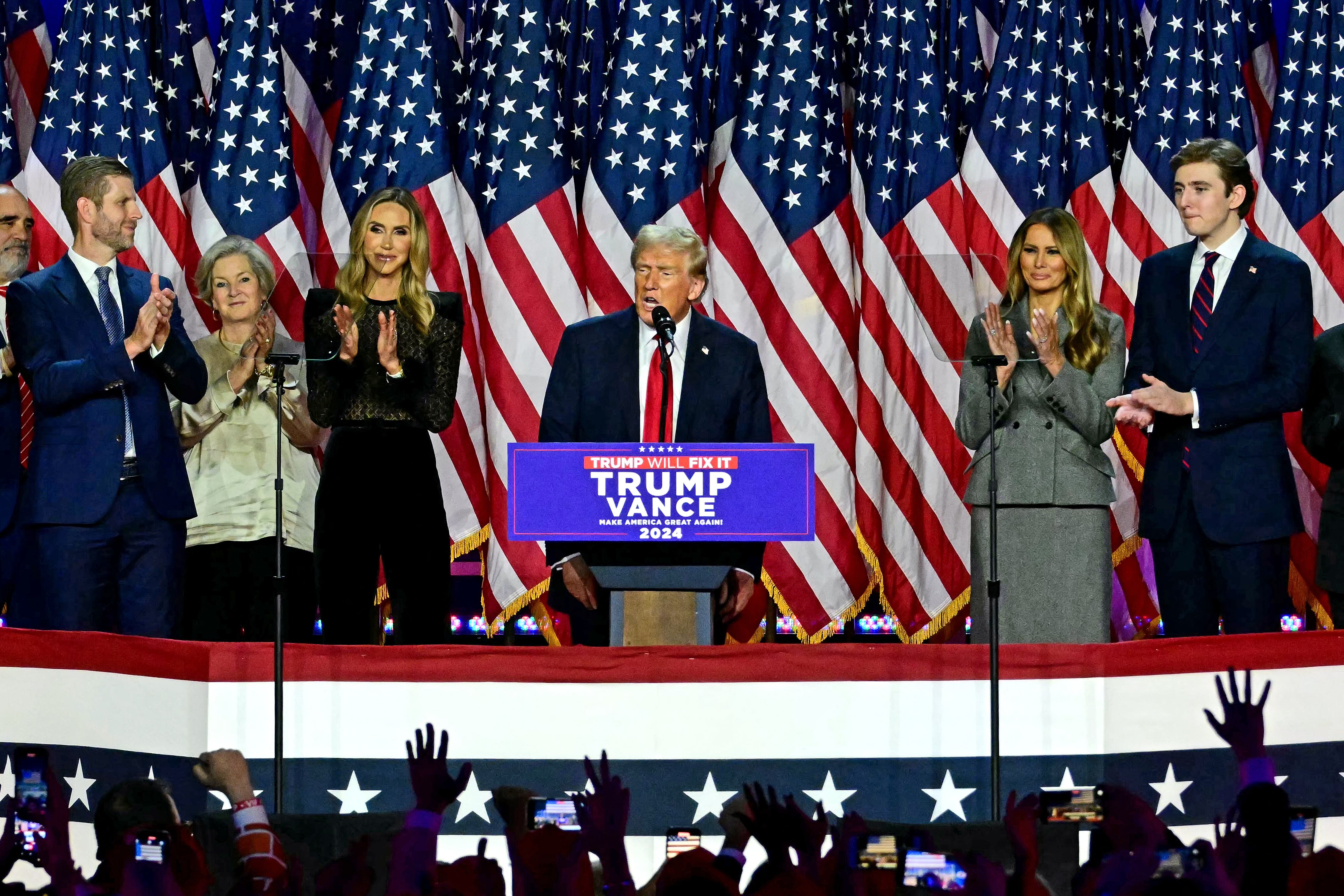 Donald Trump habla en el Centro de Convenciones de West Palm Beach rodeado de banderas de Estados Unidos tras las elecciones de 2024, acompañado de su equipo. El equipo de transición de Trump confirmó amenazas contra varios miembros del gabinete designado.