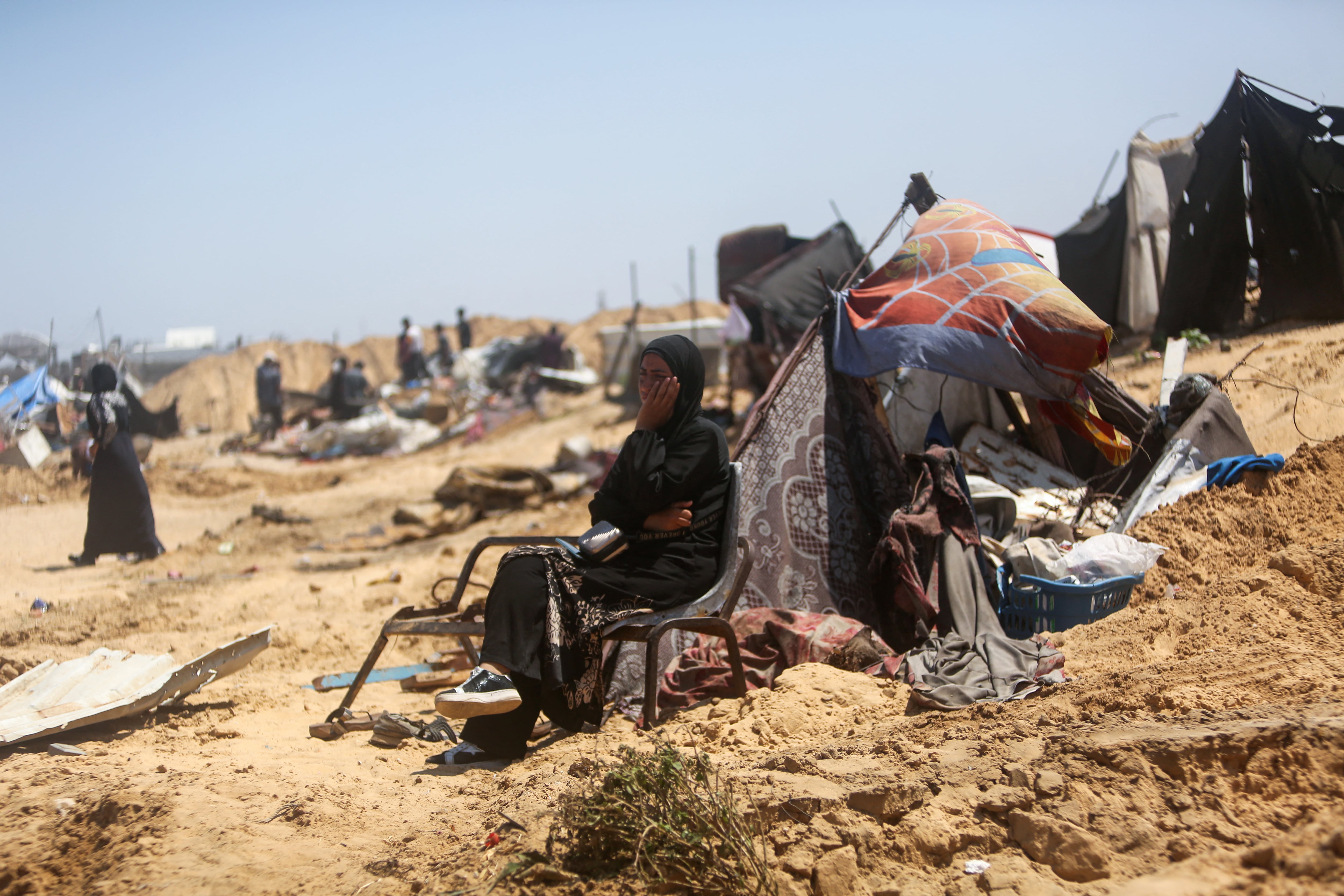 Una mujer sentada junto a sus pertenencias rescatadas observa cómo los palestinos inspeccionan la destrucción en un campamento improvisado para desplazados tras una supuesta incursión de tanques israelíes en la zona de Khan Yunis, en el sur de la Franja de Gaza, este 11 de julio. Fotografía: