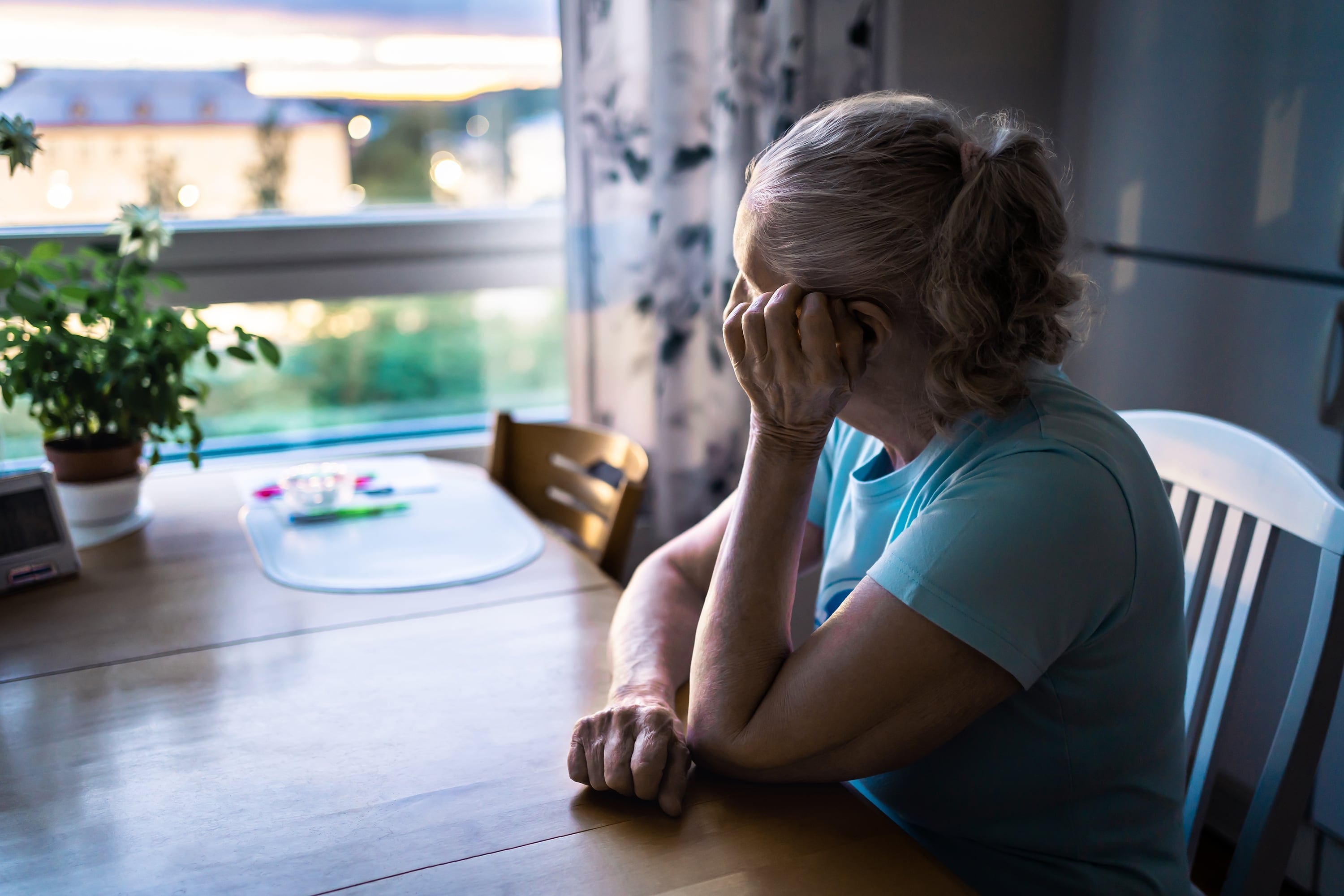 Mujer adulta mayor sentada en una mesa, reflexionando mientras mira por la ventana, símbolo de la importancia de la compañía en la tercera edad.