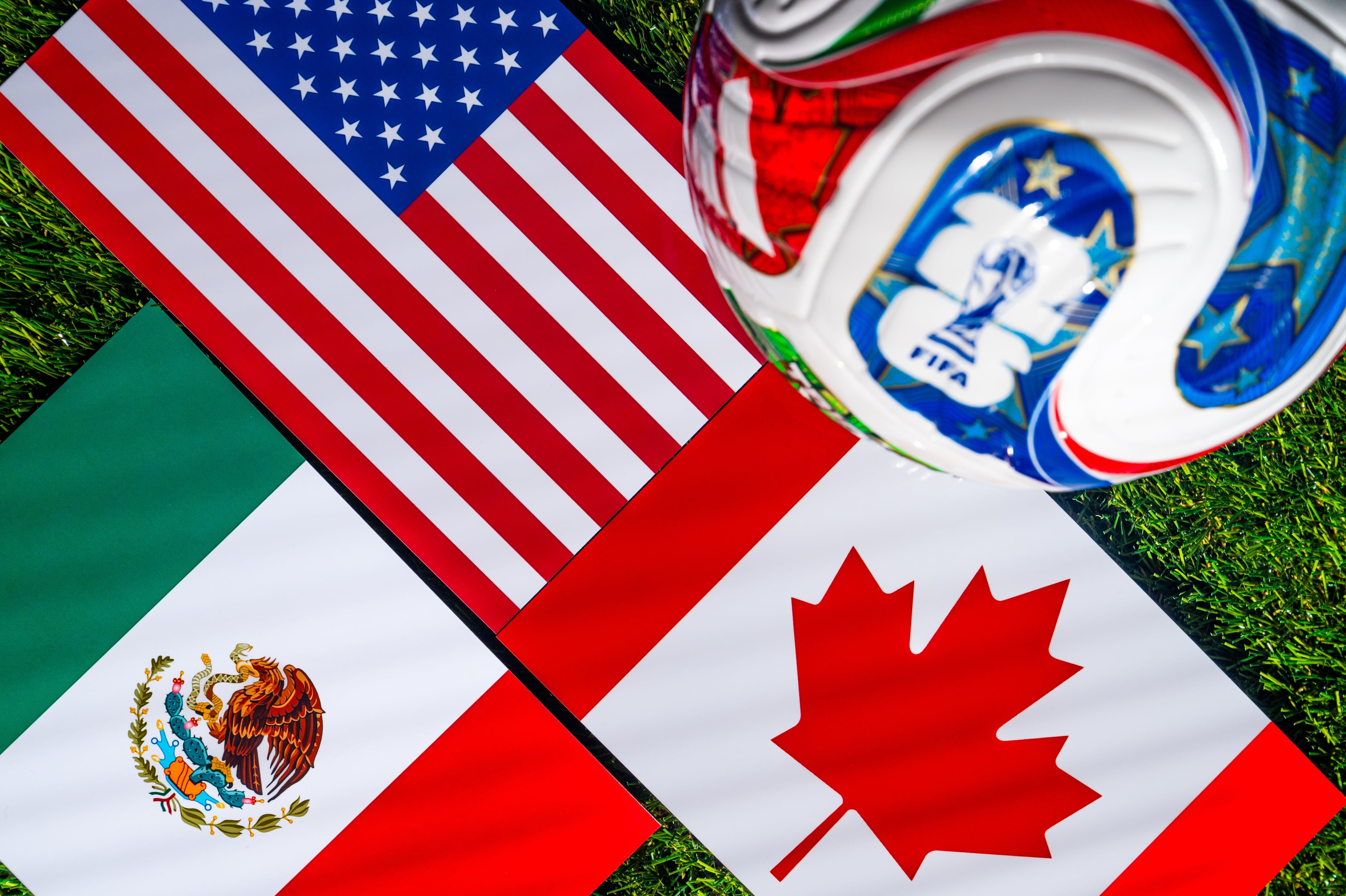 Banderas de Estados Unidos, Canadá y México, países anfitriones del Mundial 2026, junto a un balón de fútbol sobre el césped