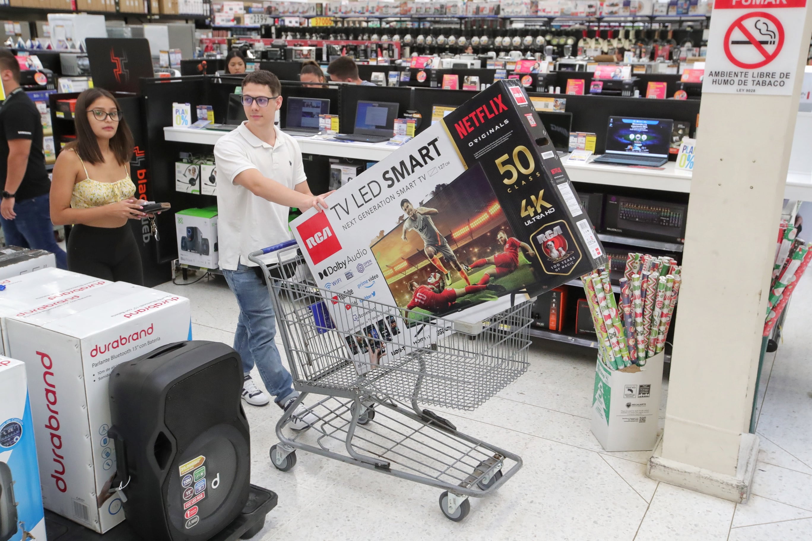 Un hombre empuja un carrito de compras con un televisor 4K mientras una mujer lo acompaña en una tienda departamental, rodeados de productos electrónicos.