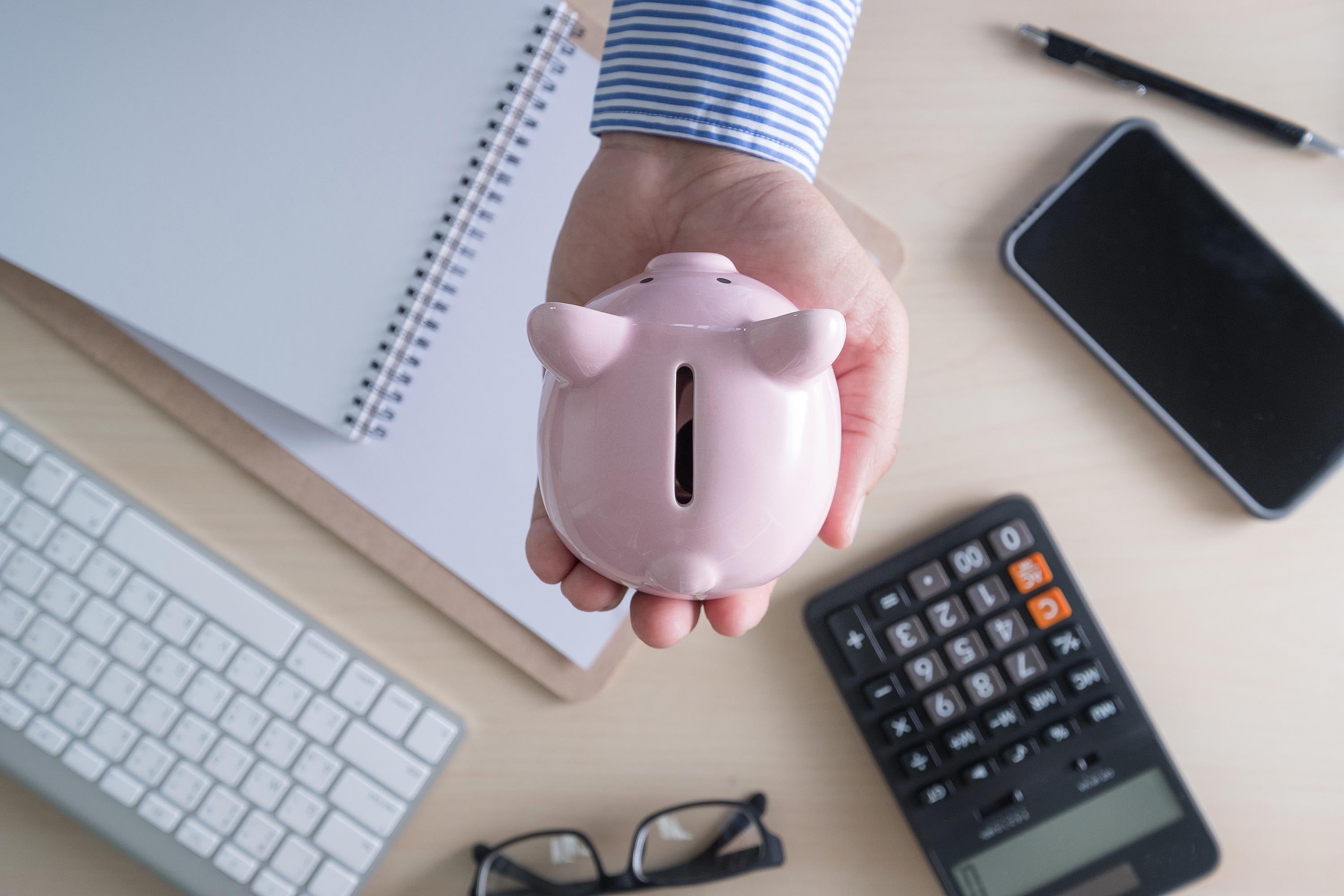 Primer plano de una mano sosteniendo una alcancía rosa sobre un escritorio con calculadora, libreta, teclado y celular, simbolizando el ahorro y la planificación financiera.