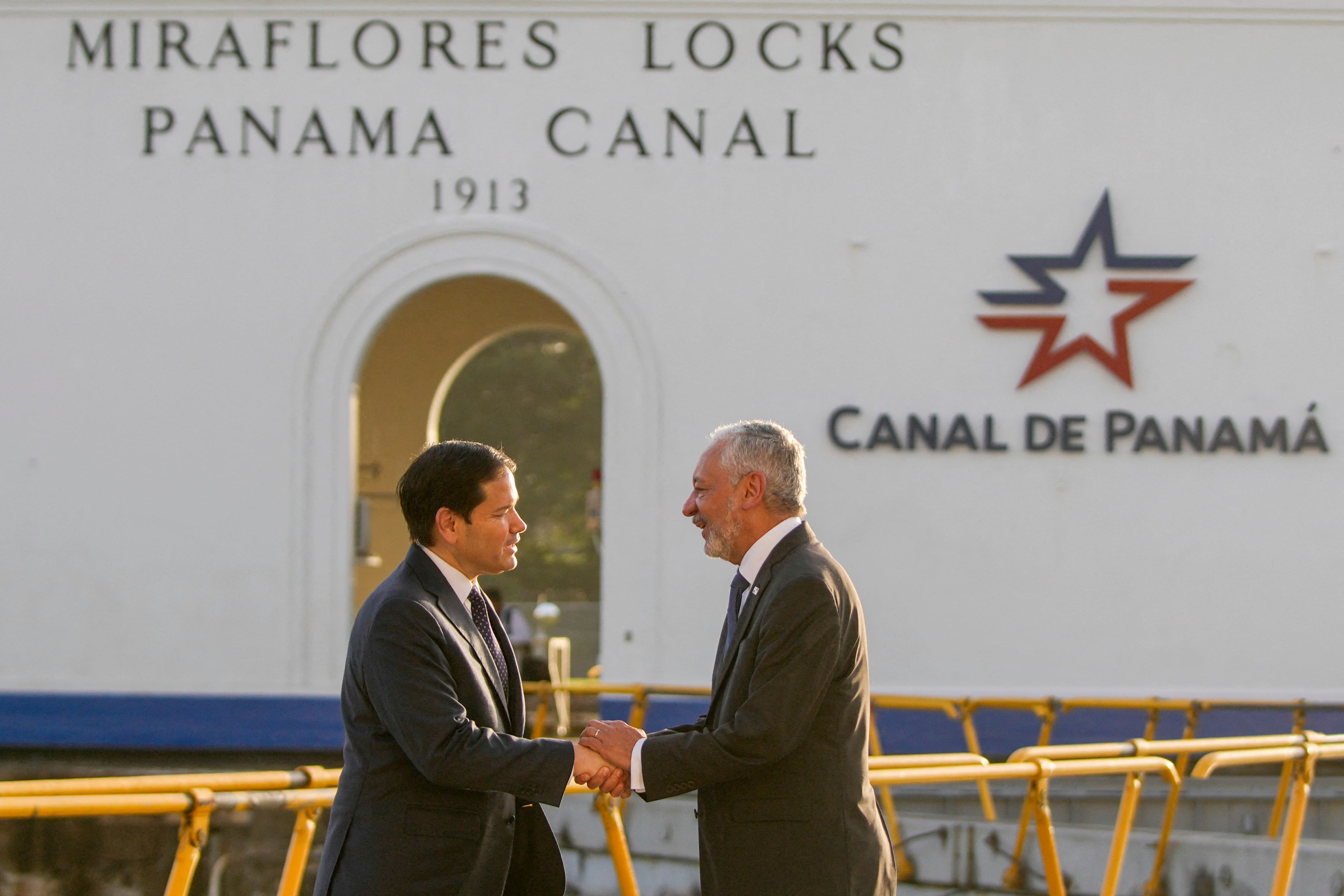 Dos hombres se dan la mano en señal de saludo frente a la entrada principal del Canal de Panamá. Visten traje entero.