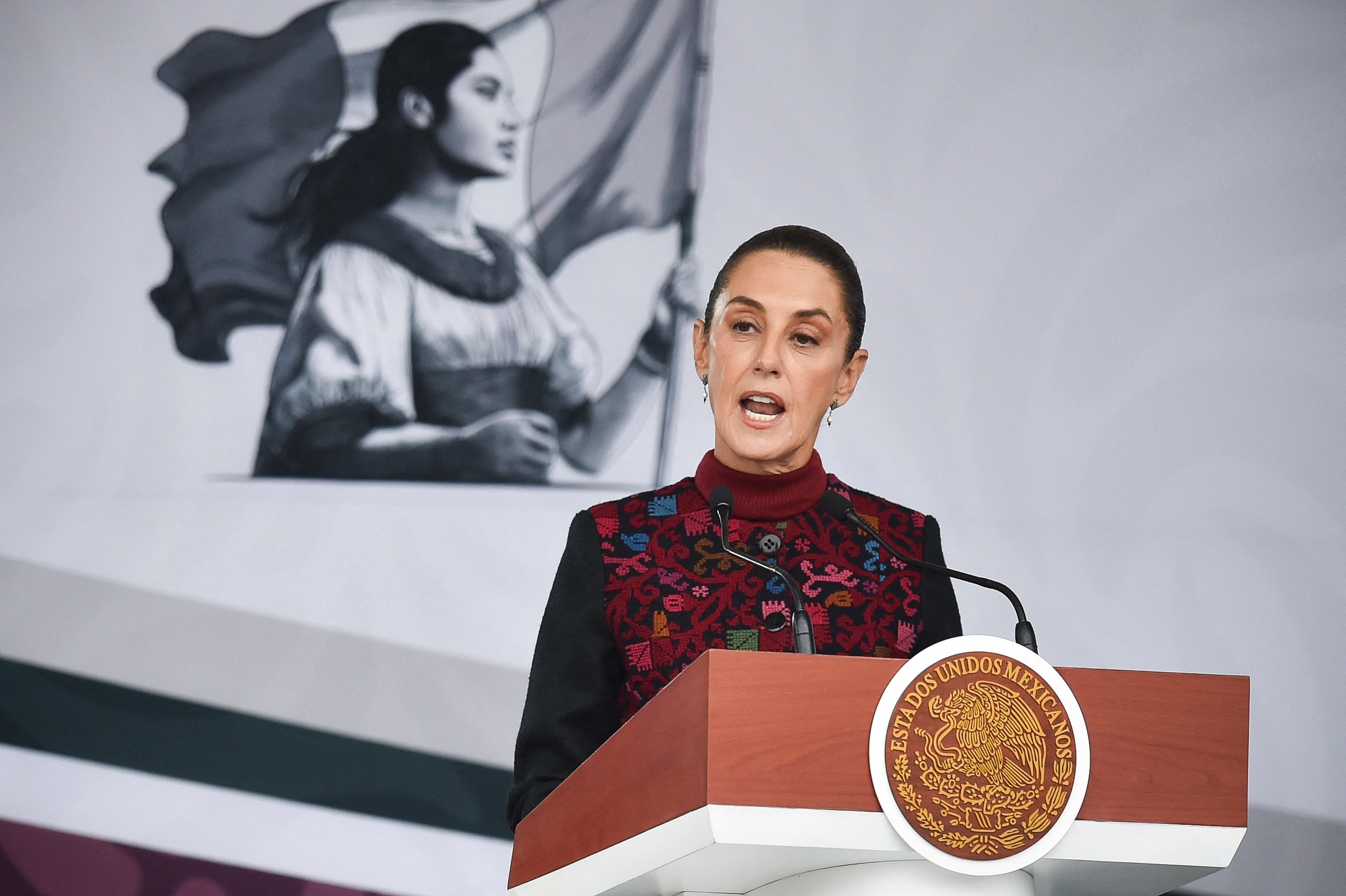La presidenta de México, Claudia Sheinbaum, dando un discurso frente a un podio, con un mural patriótico de una mujer con una bandera en el fondo.