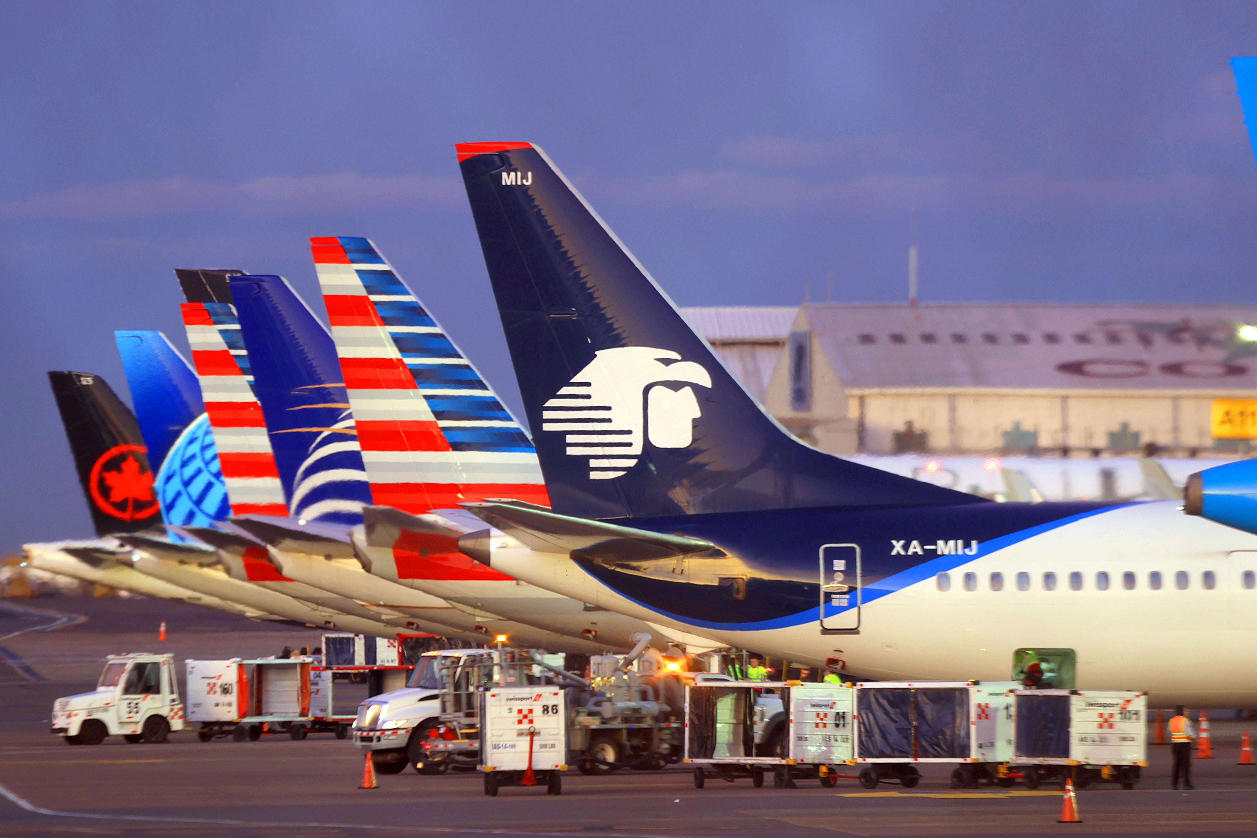 Filas de aviones de distintas aerolíneas estacionados en el Aeropuerto Juan Santamaría al atardecer, con equipos de carga operando en la pista.
