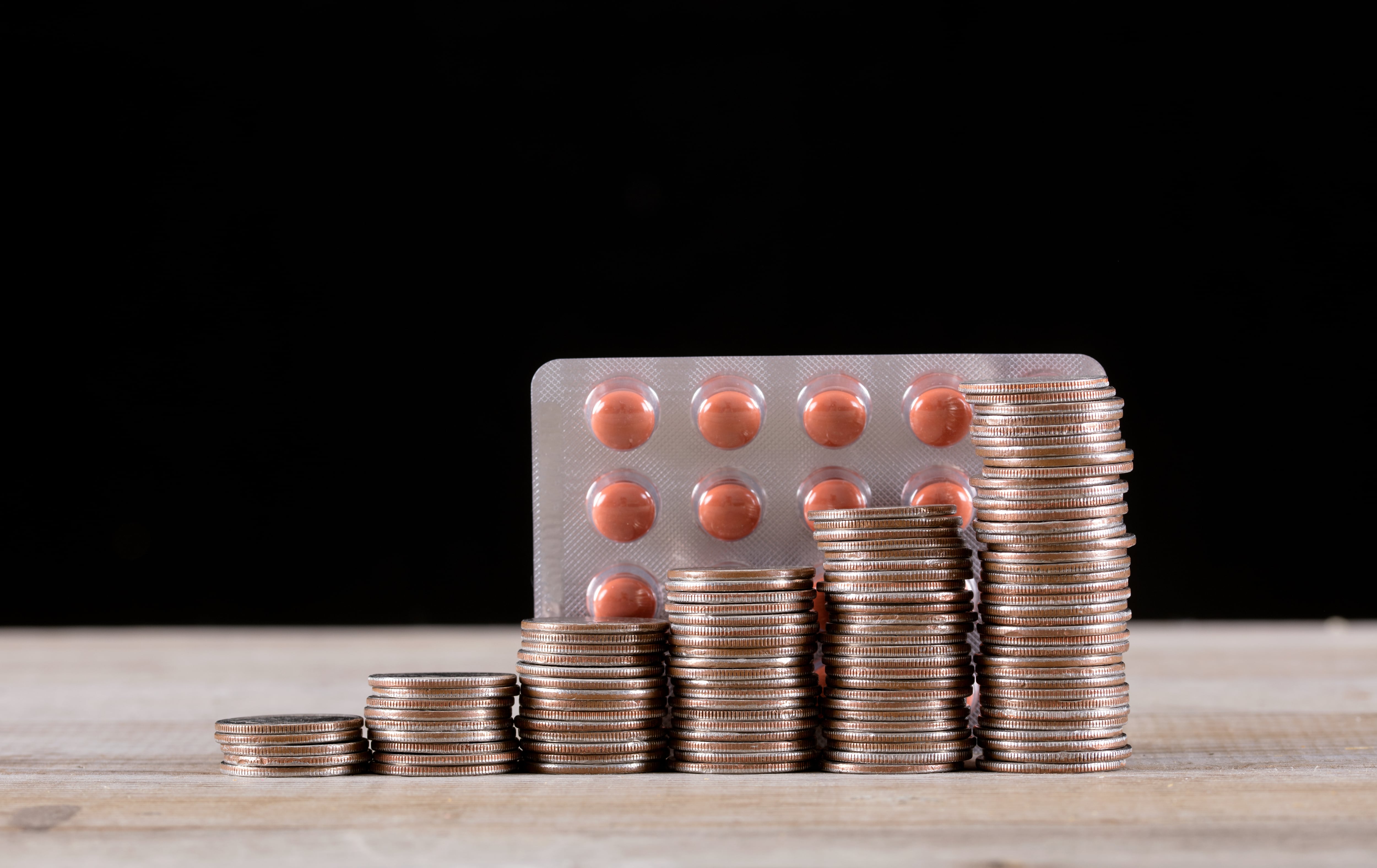 Blister con pastillas anaranjadas, fondo negro, sobre mesa café claro. Al frente del blister, torre de monedas.