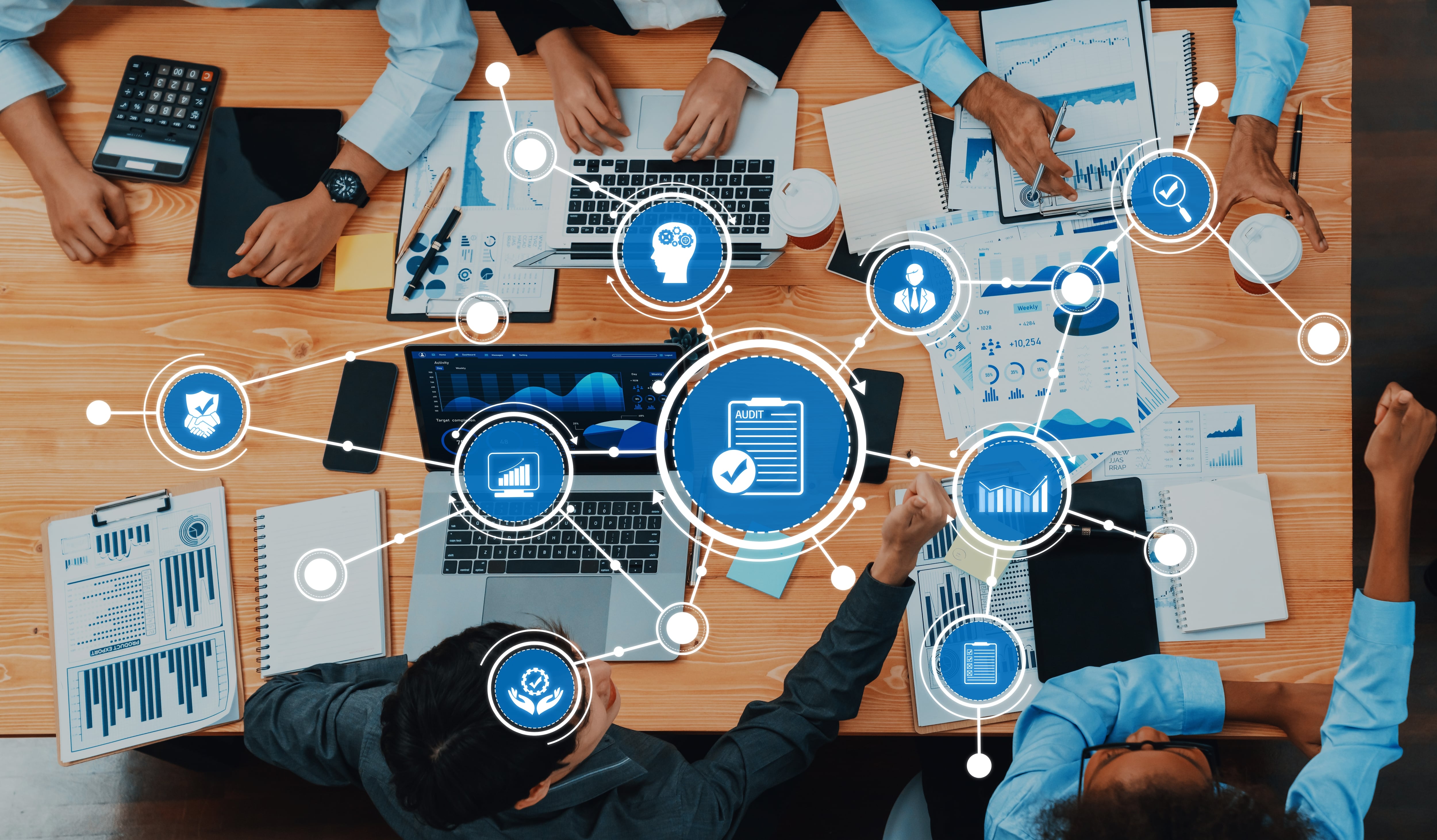 Gobernanza con inteligencia artificial o IA. Un espacio de trabajo dinámico que muestra la colaboración con tecnología y análisis. Muestra manos sobre una mesa con computadoras portátiles, documentos y gráficos de datos en un entorno empresarial.