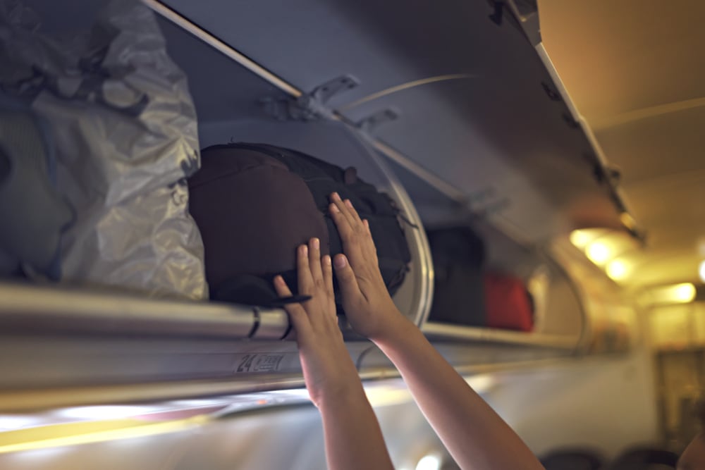 Manos de un pasajero acomodando su equipaje de mano en el compartimento superior de un avión. Las aerolíneas aplican diferentes reglas sobre el tamaño y peso del equipaje de cabina.