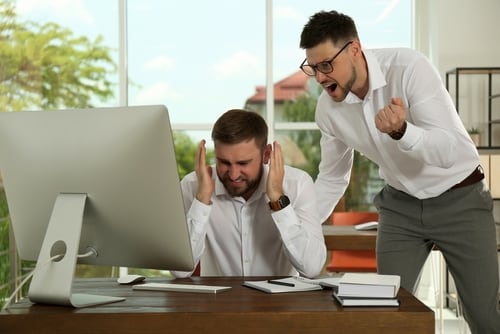 Un hombre joven se lleva las manos a la cabeza frente a una computadora mientras otro hombre lo señala y grita, ilustrando el acoso laboral en el lugar de trabajo.