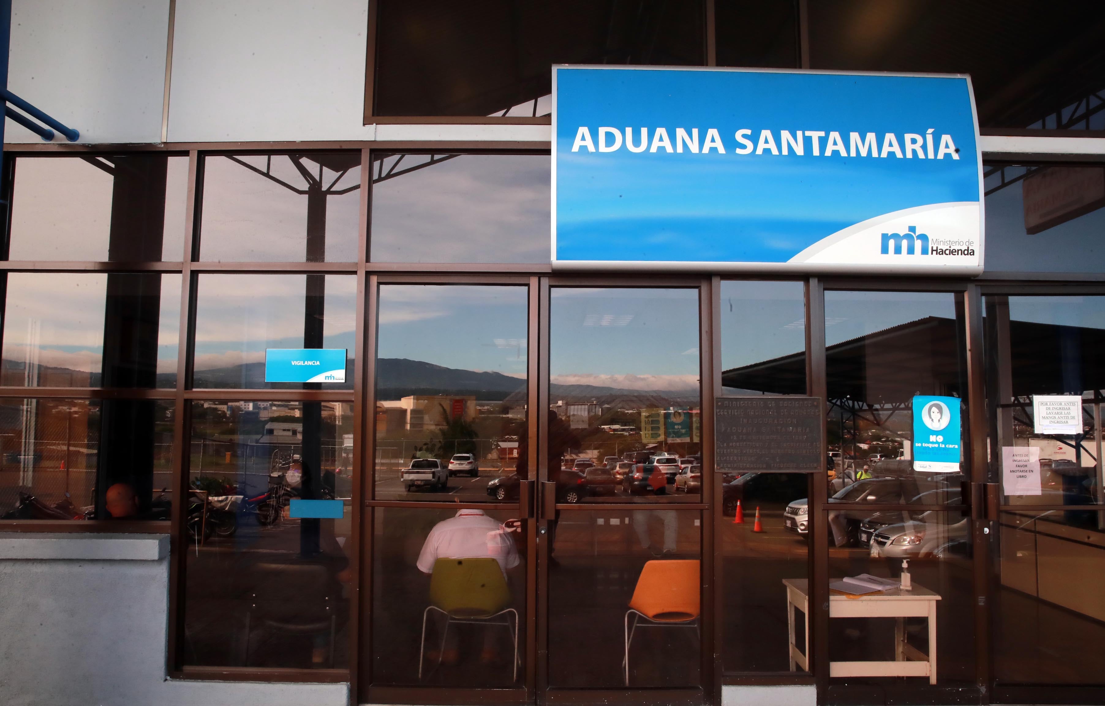 Vista frontal de la entrada de la Aduana Santamaría en Costa Rica, con reflejos del paisaje urbano y montañas en los vidrios. Este edificio está asociado a los servicios del Ministerio de Hacienda y la plataforma ‘Aduana Fácil’.