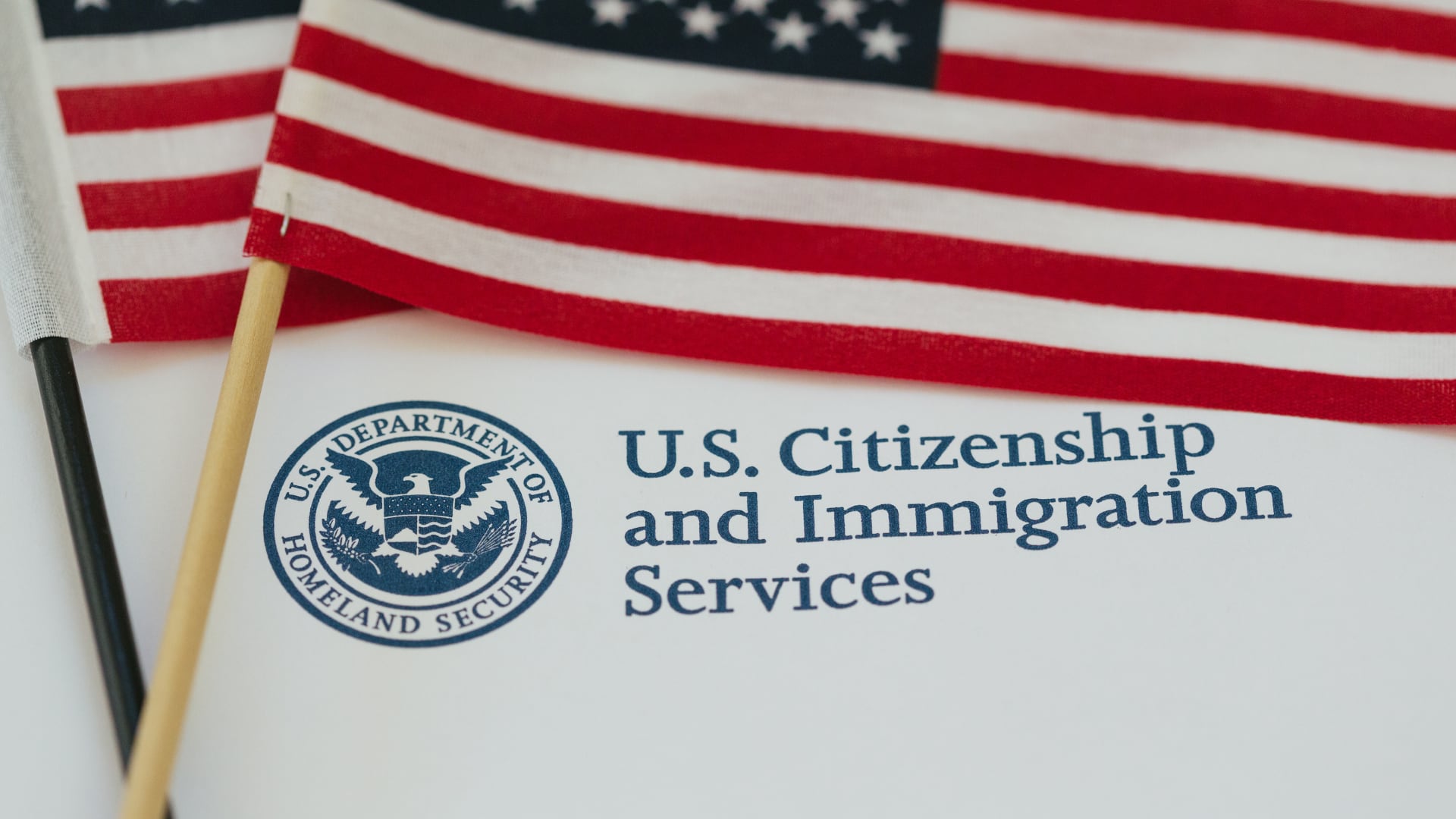 Logo del Servicio de Ciudadanía e Inmigración de Estados Unidos (USCIS) junto a una bandera de Estados Unidos, en el contexto de nuevas medidas para exigir datos biométricos a personas migrantes registradas.