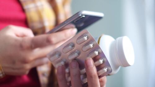 Mujer sosteniendo medicamentos y usando un teléfono móvil para comparar precios de medicinas.