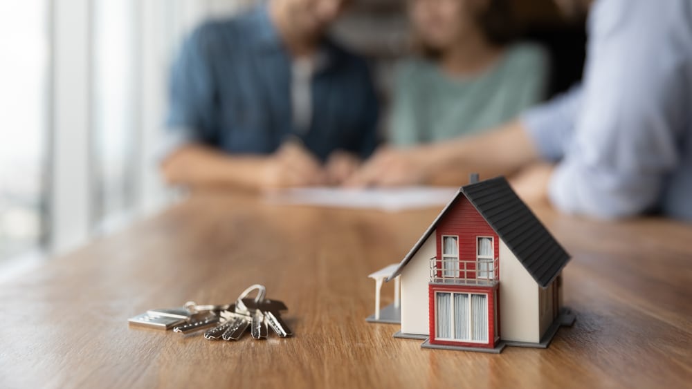 Primer plano de una casa a escala y un juego de llaves sobre una mesa, con una pareja difuminada firmando documentos al fondo. Representa bienes inmuebles y el contexto del impuesto sobre bienes inmuebles.
