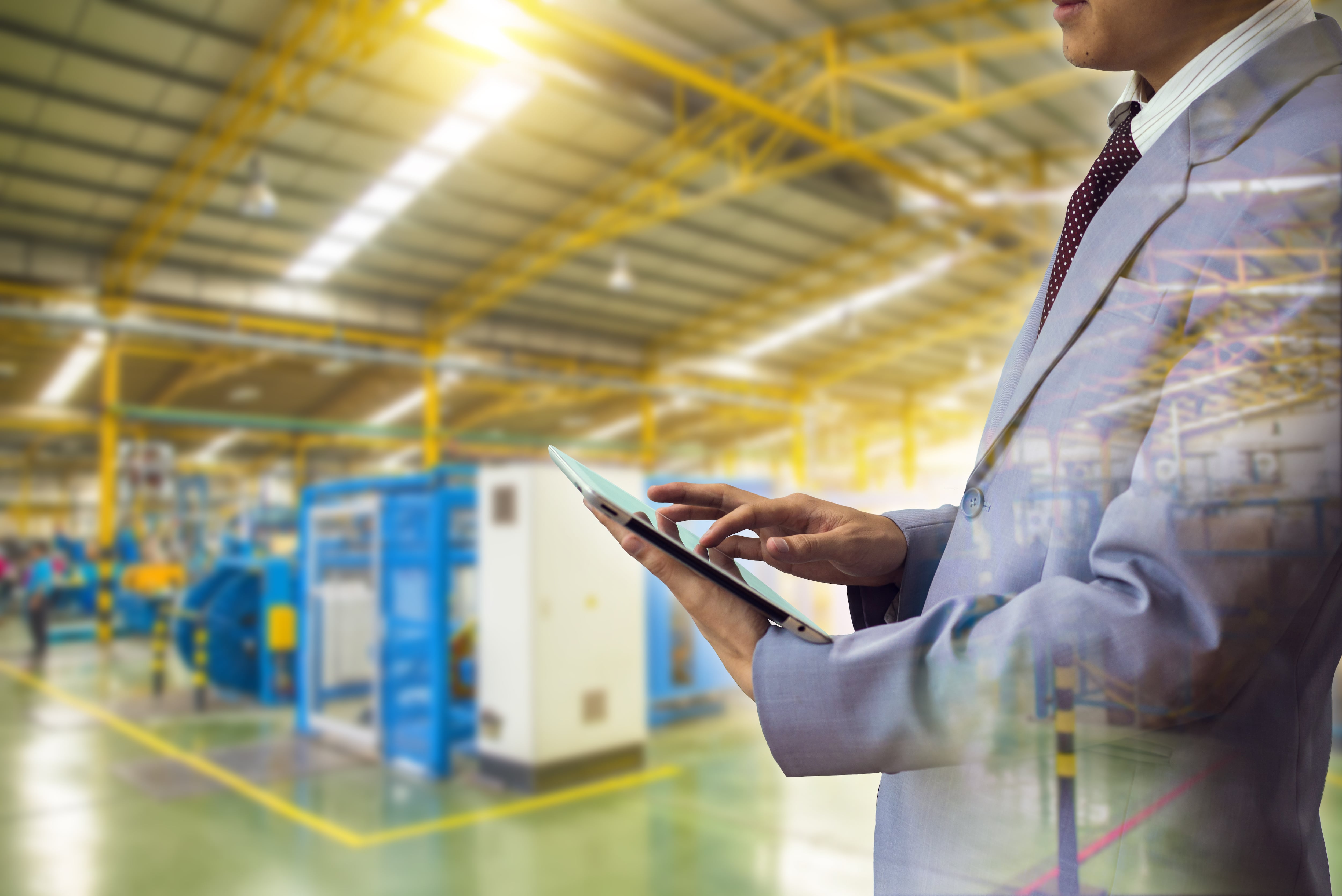 Planta de manufactura en segundo plano y un hombre con traje sostiene una tablet.