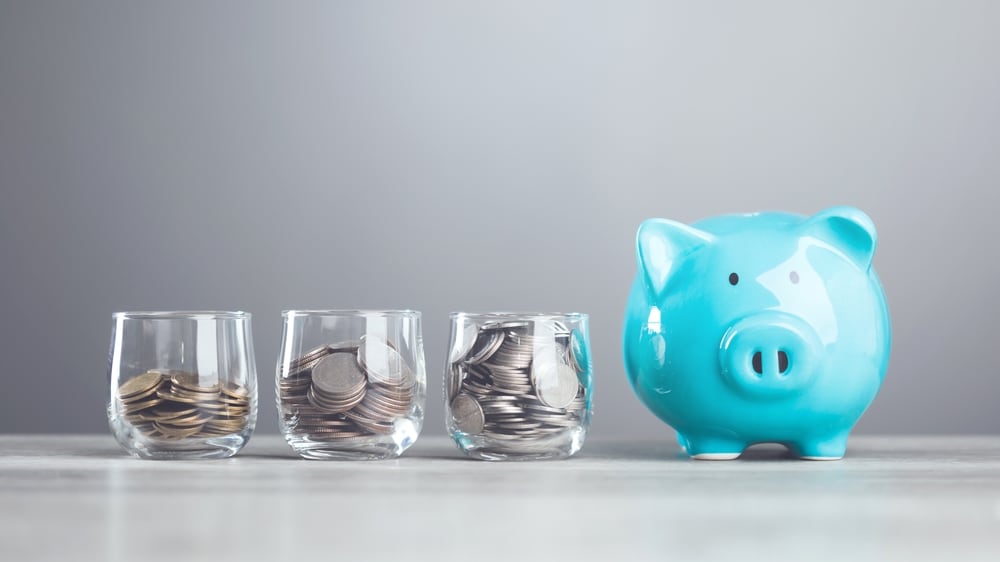 Alcancía azul junto a tres vasos con monedas que representan opciones de inversión y diversificación de capital.