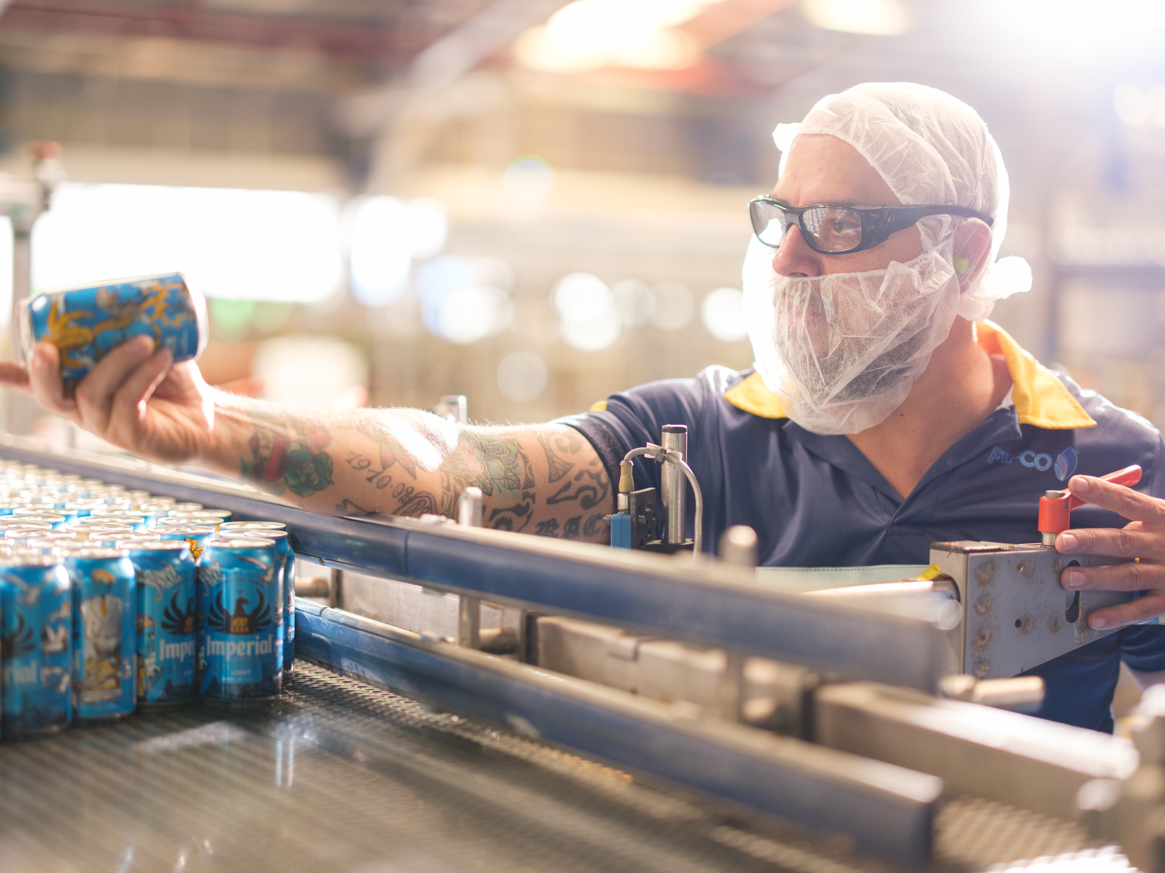 Trabajador con malla de protección y gafas inspeccionando una lata de cerveza en la línea de producción.