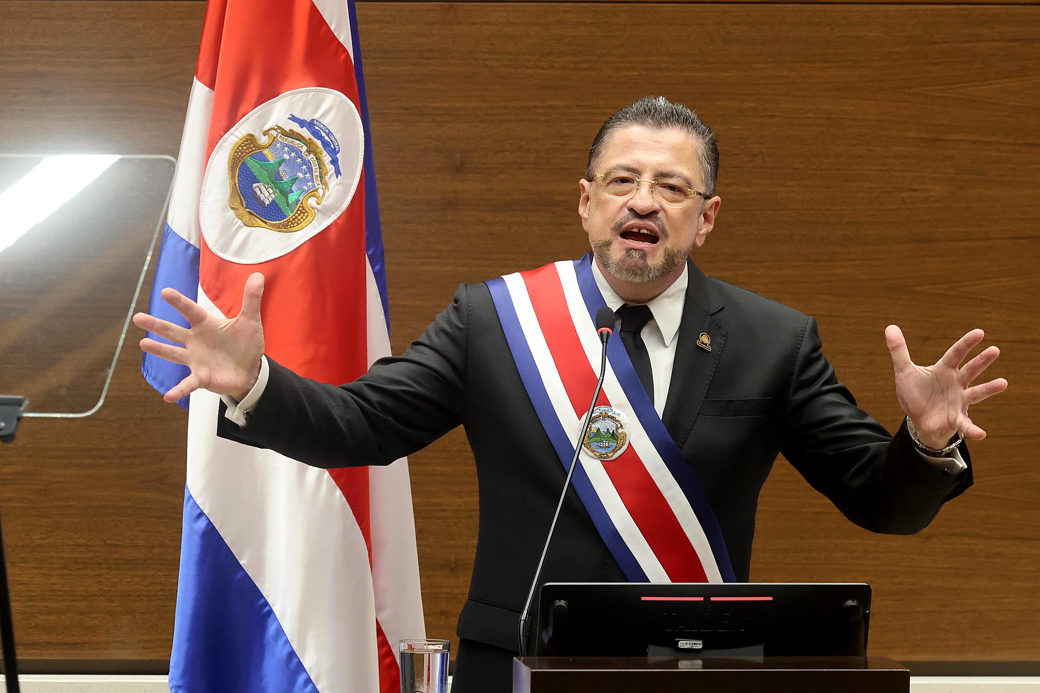 El presidente de Costa Rica, con banda presidencial, ofrece un discurso en un acto oficial con la bandera de Costa Rica al fondo.