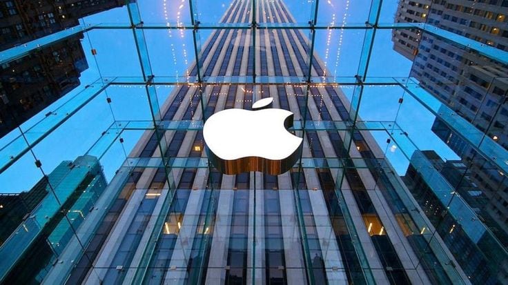 Fachada de cristal de una tienda Apple con el logo de la marca iluminado, reflejando rascacielos.