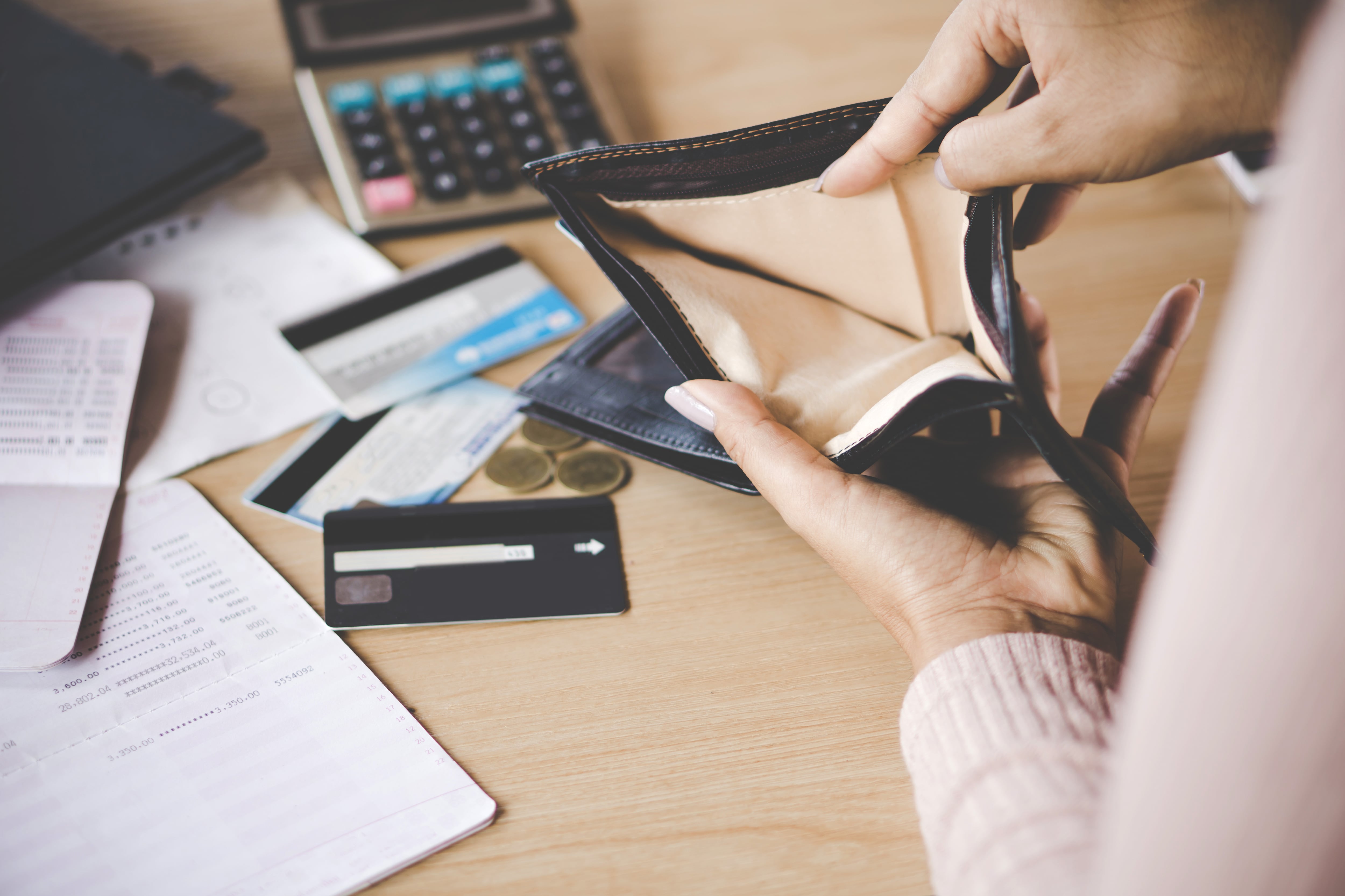 Una persona muestra su billetera sin dinero. En la mesa hay tarjetas de créditos, monedas, papeles y una calculadora.