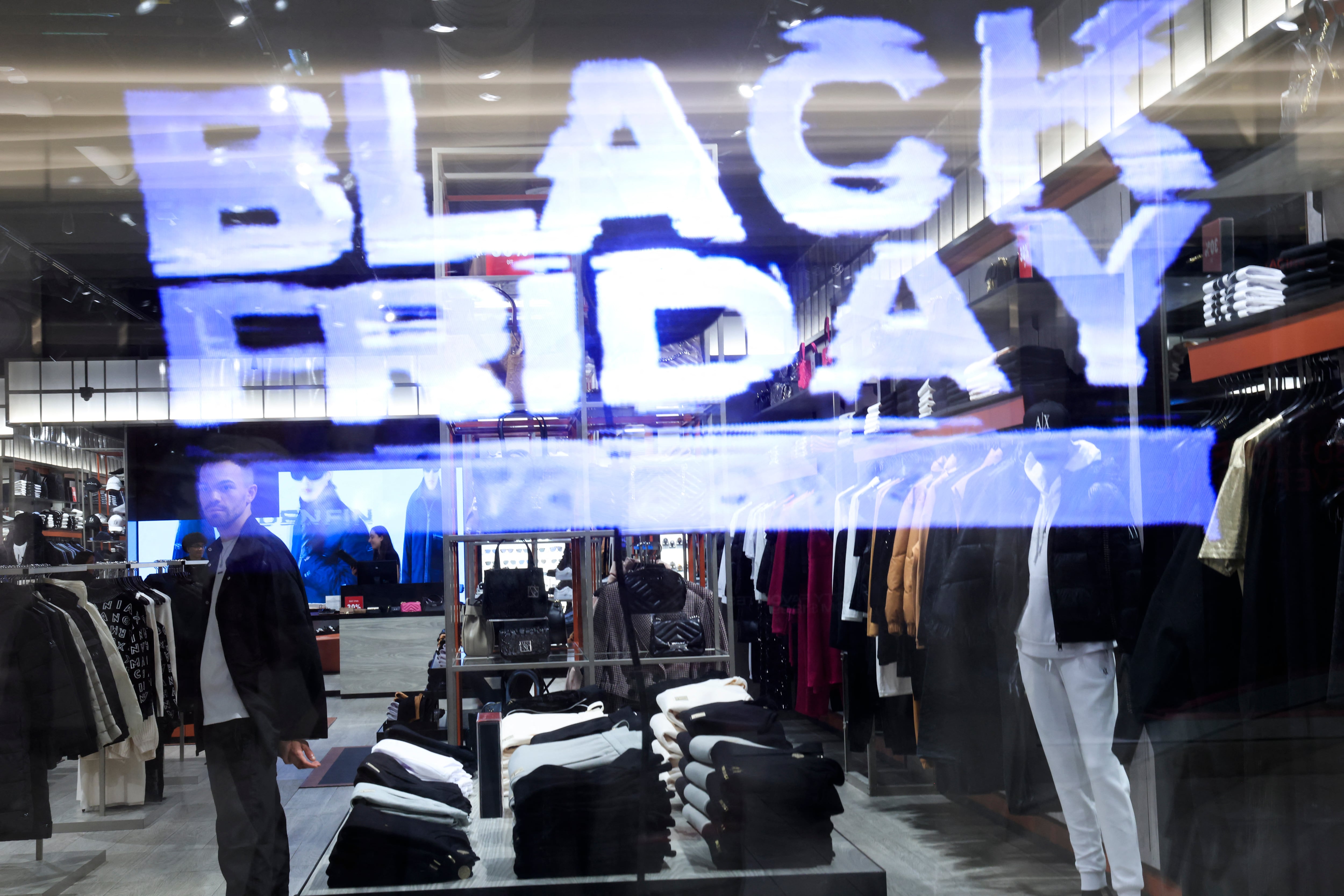 Vista de una tienda de ropa con la palabra 'Black Friday' reflejada en el vidrio, mientras un cliente recorre los estantes en busca de ofertas.