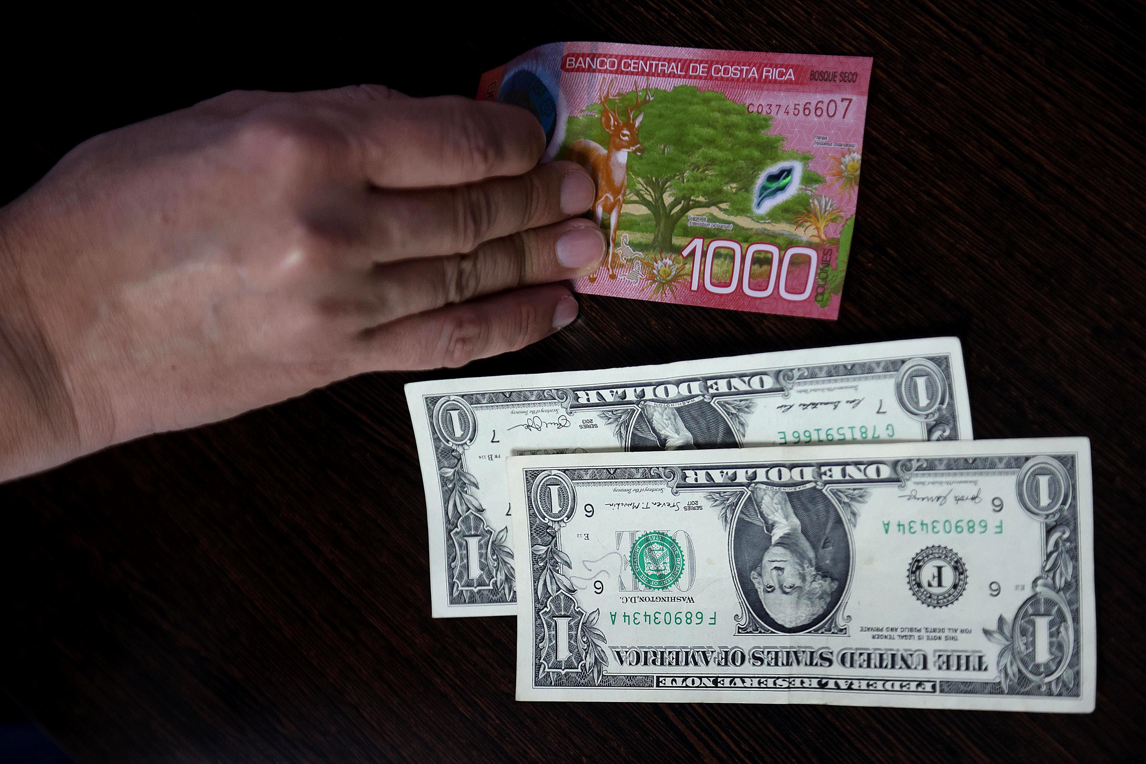 Mano sosteniendo un billete de ¢1.000 junto a dos billetes de $1, reflejando la variación en el tipo de cambio reportada en el Mercado de Monedas Extranjeras (Monex) en Costa Rica.