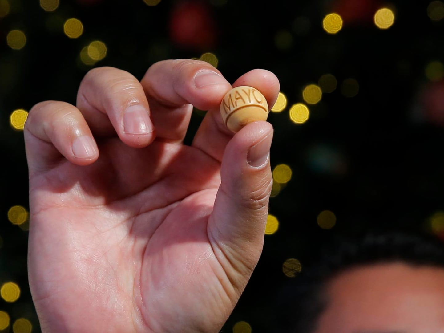 Mano sosteniendo una bola de sorteo del Gordo Navideño 2023 marcada con el mes de mayo, sobre un fondo decorativo con luces desenfocadas.