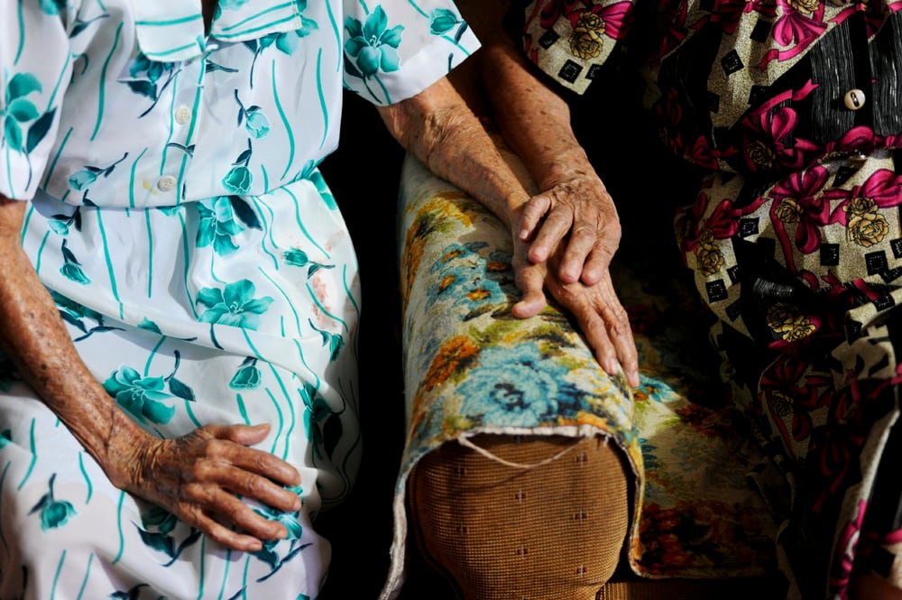 Las manos y cuerpos de dos adultas mayores. Están juntas, una tiene su mano sobre la mano de la otra.