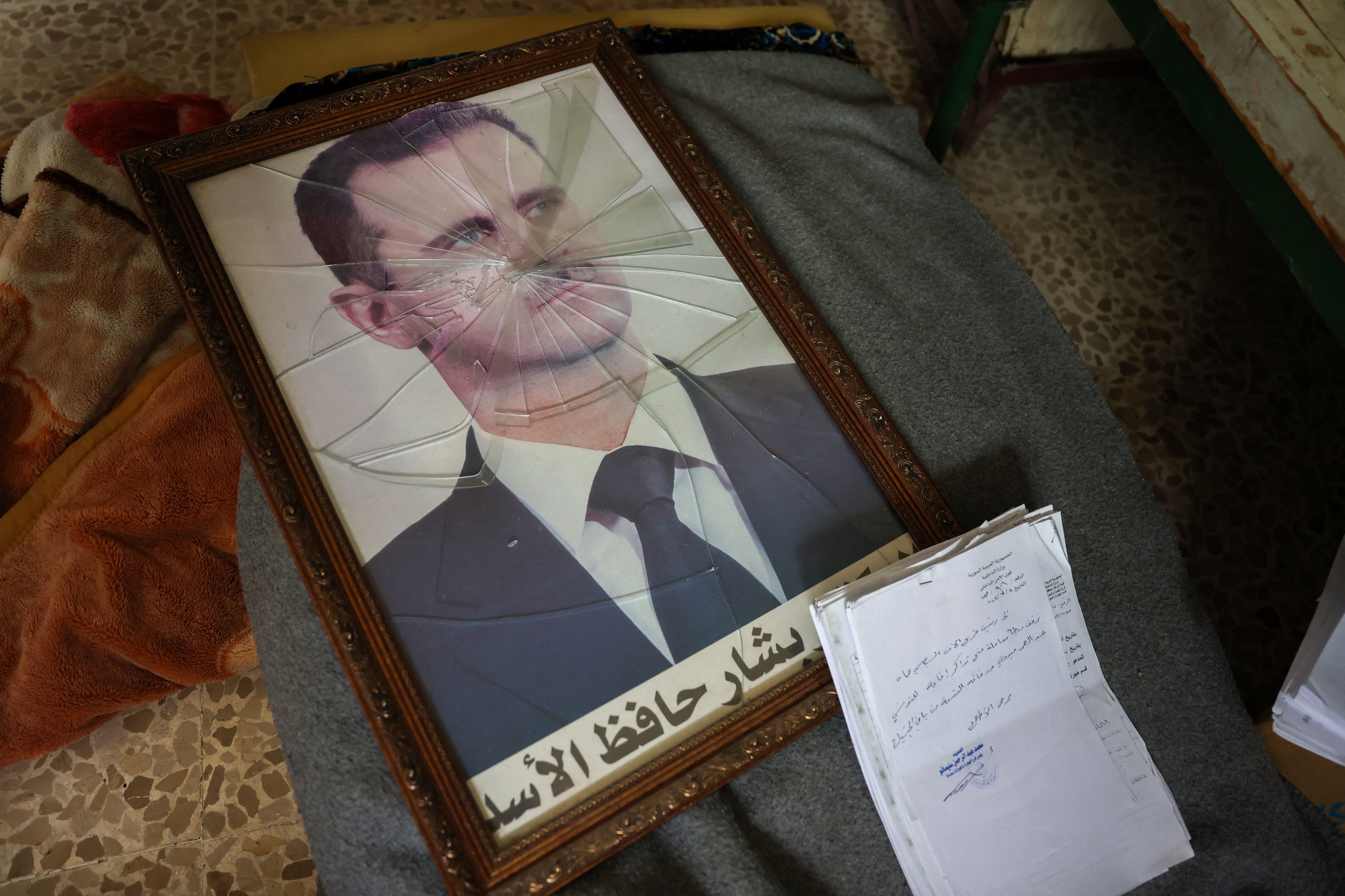 Retrato quebrado de Bashar al Asad con vidrio roto sobre una mesa junto a documentos, simbolizando el colapso de su régimen en Siria.
