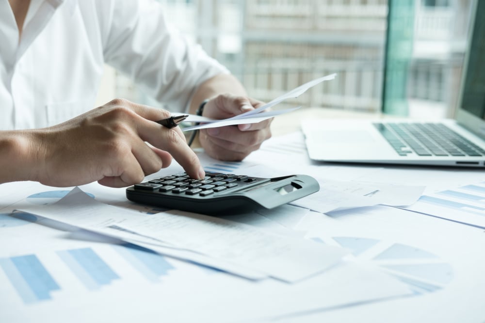 Manos de una persona usando una calculadora y revisando documentos financieros sobre una mesa, con gráficos y una computadora portátil, representando la preparación de la declaración de renta 2024.