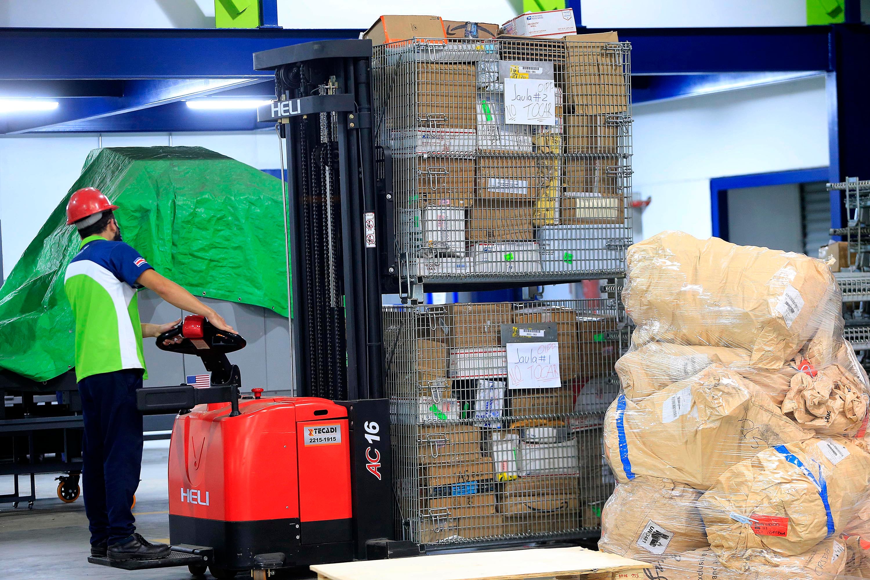 Un operario en una bodega manipula paquetes con un montacargas eléctrico. Los paquetes están organizados en jaulas metálicas y bolsas para distribución.